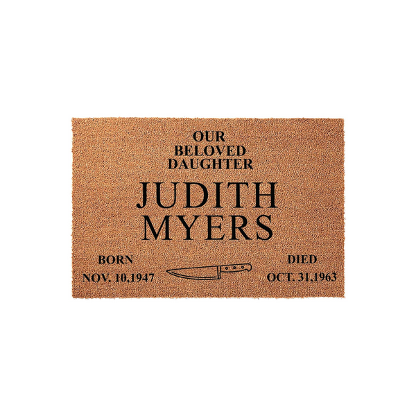 Horror movie themed doormat featuring a natural coir texture with black printed text that reads Our Beloved Daughter Judith Myers, including birth and death dates and a graphic of a kitchen knife centered on a plain white studio