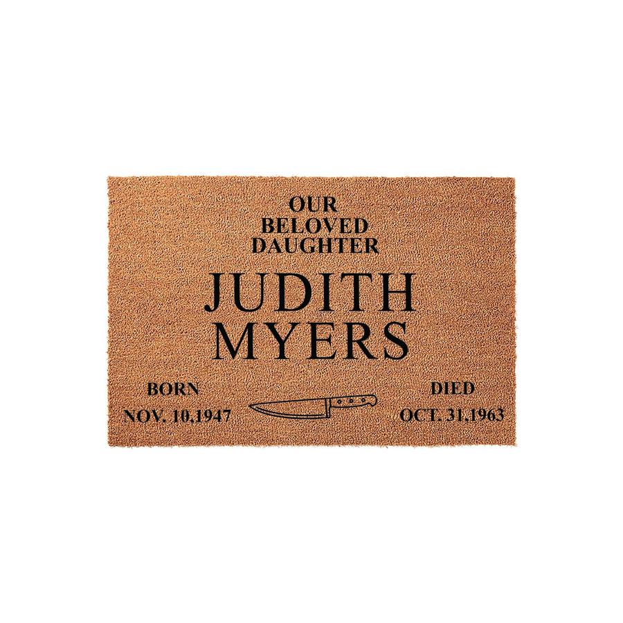 Horror movie themed doormat featuring a natural coir texture with black printed text that reads Our Beloved Daughter Judith Myers, including birth and death dates and a graphic of a kitchen knife centered on a plain white studio