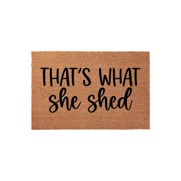 That's What She Shed coir doormat featuring a natural tan fiber texture and bold black lettering, designed as a humorous outdoor welcome mat for a backyard garden shed or a creative hobby space on a plain white studio background.