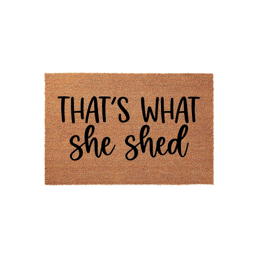 That's What She Shed coir doormat featuring a natural tan fiber texture and bold black lettering, designed as a humorous outdoor welcome mat for a backyard garden shed or a creative hobby space on a plain white studio background.