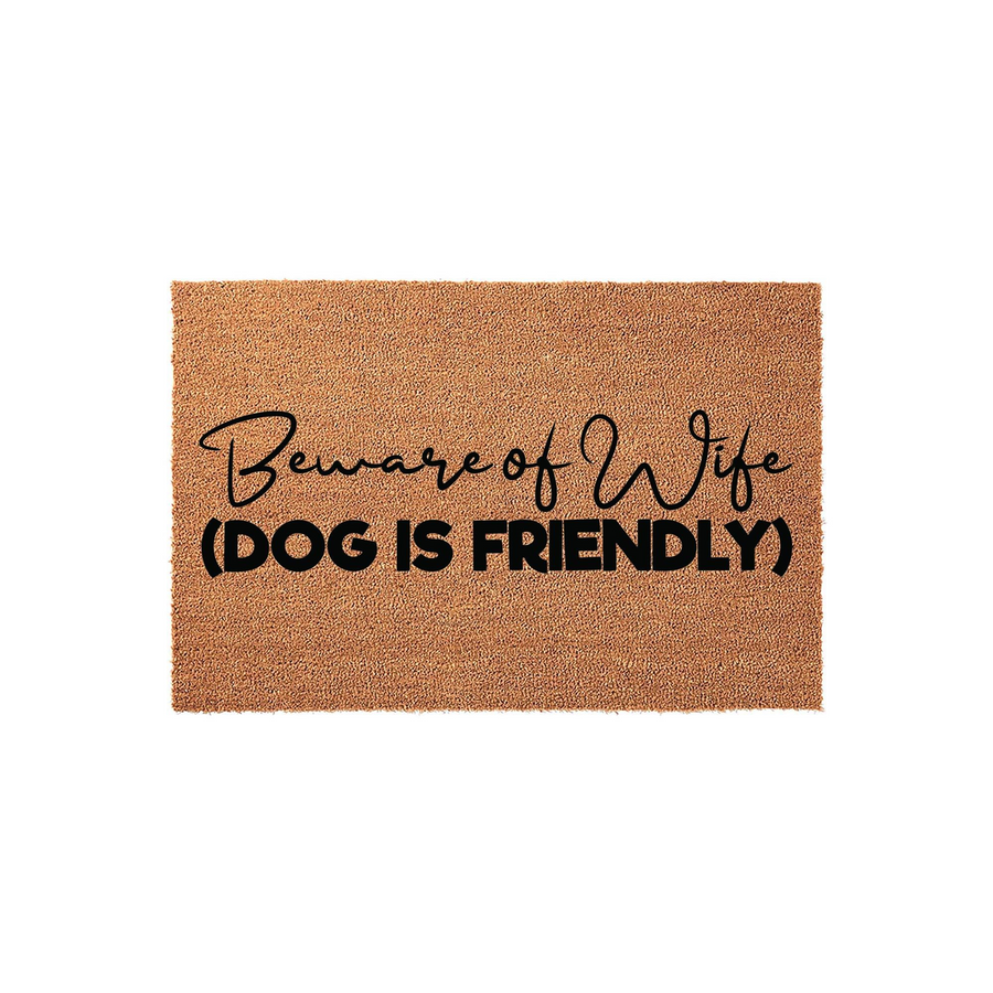 Funny Beware of Wife Dog Is Friendly doormat made of natural coir fiber, featuring bold black text in a mix of script and block fonts centered on a rectangular brown mat against a clean white studio background for a humorous home
