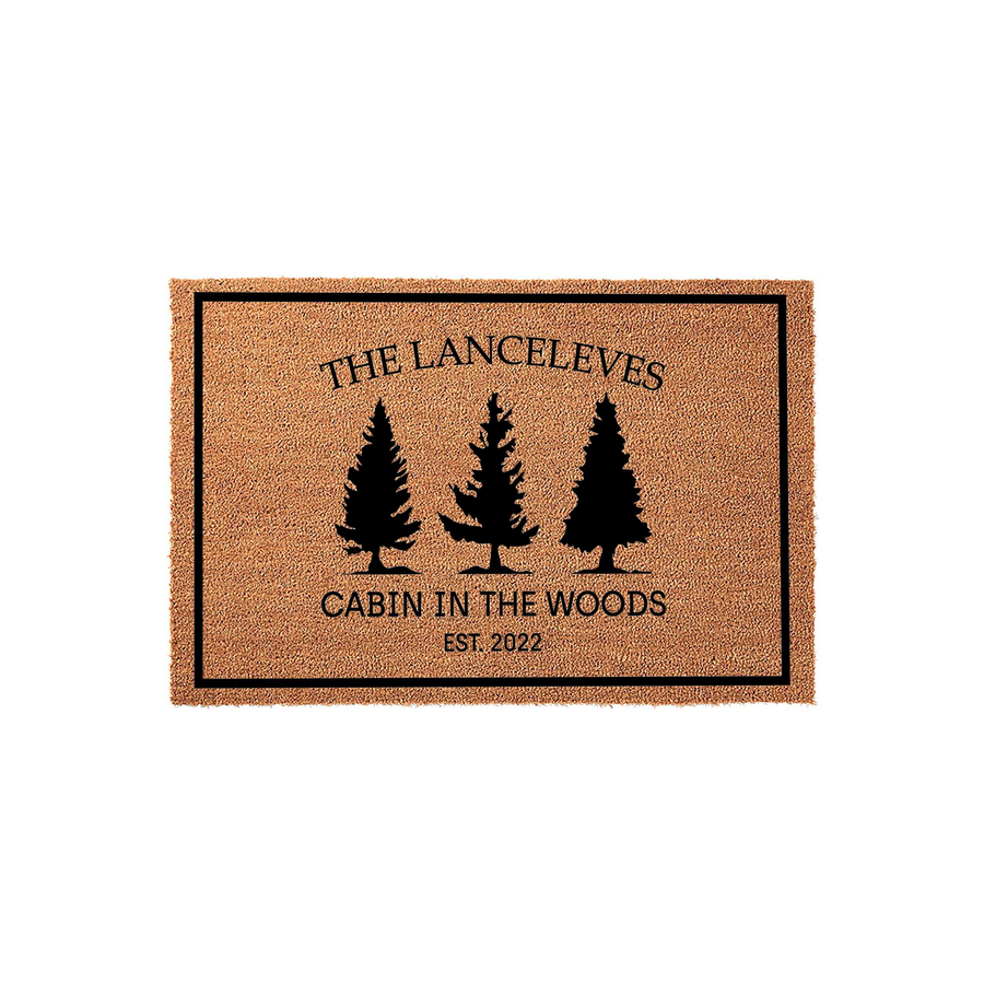Personalized Cabin in the Woods doormat featuring a natural coir texture, black pine tree silhouettes, and custom family name text with an established year, all framed by a simple black border on a clean white studio background.