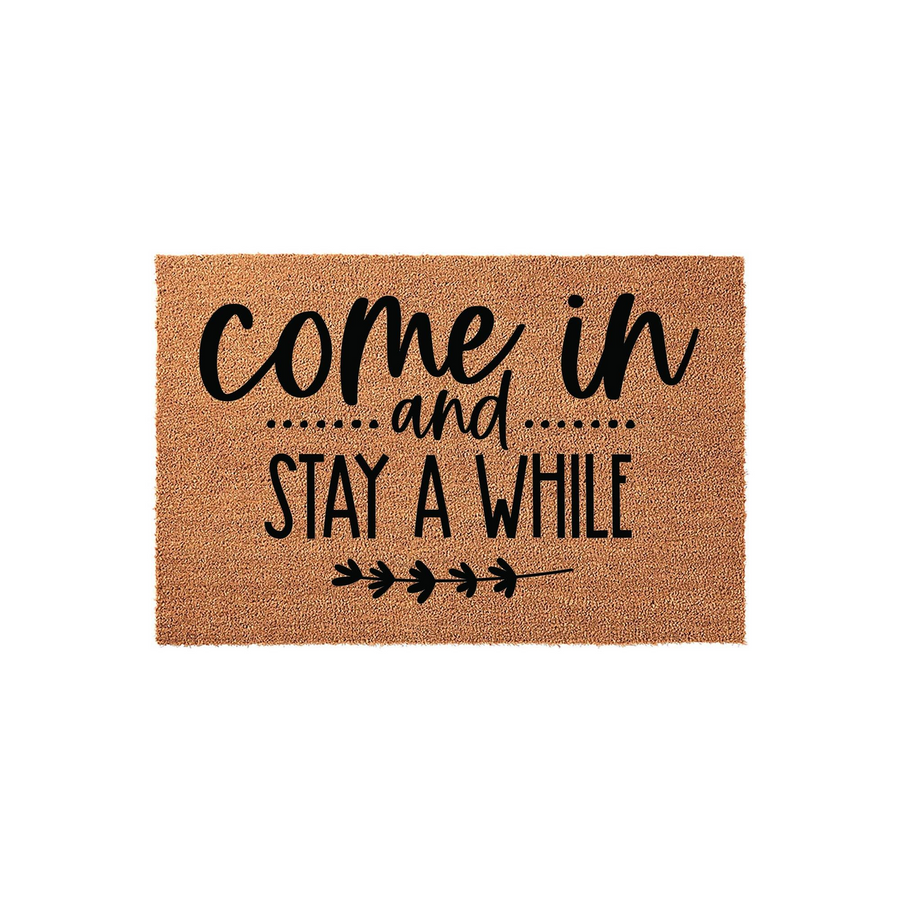 Come In and Stay A While coir doormat featuring a natural tan fiber texture and bold black typography, accented with a simple decorative leaf branch design at the bottom for a welcoming farmhouse style front porch entrance.