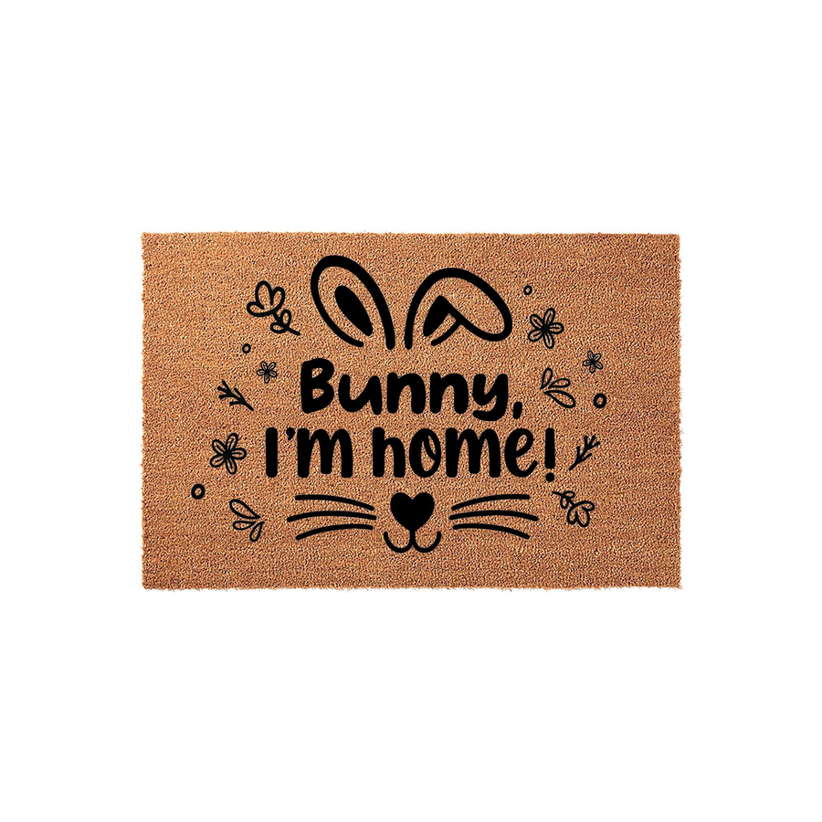 Bunny I'm Home coir doormat featuring a playful black graphic of rabbit ears, whiskers, and a heart-shaped nose surrounded by small floral accents on a natural brown textured fiber background for a festive spring entryway decor.