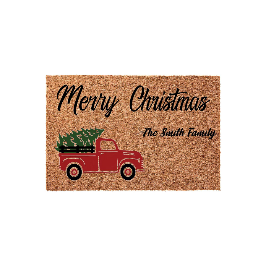 Personalized Merry Christmas coir doormat featuring a vintage red truck carrying a green pine tree, with custom family name text printed in black script on a natural tan textured background for a festive holiday home entrance