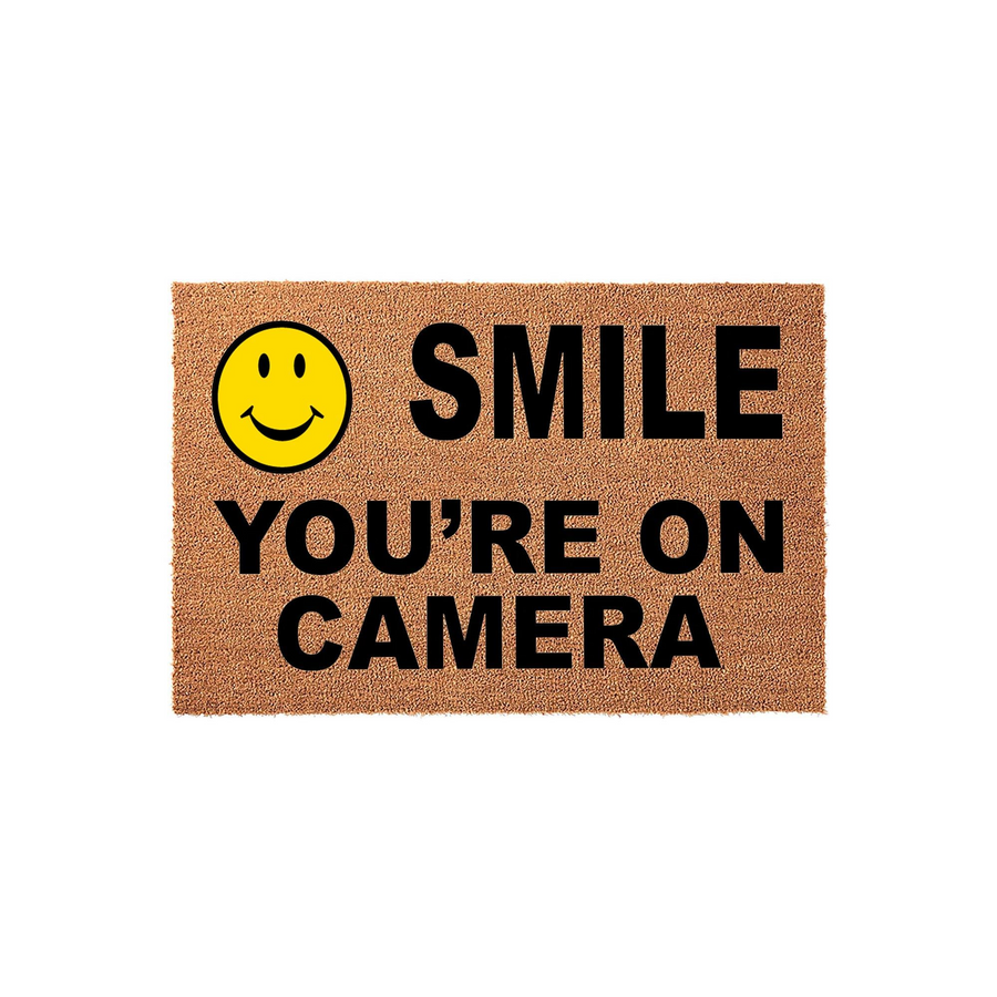 Smile You Are On Camera doormat featuring a bright yellow smiley face icon and bold black text printed on a natural brown coir fiber mat, providing a humorous and functional welcome for your front porch or entryway home decor.