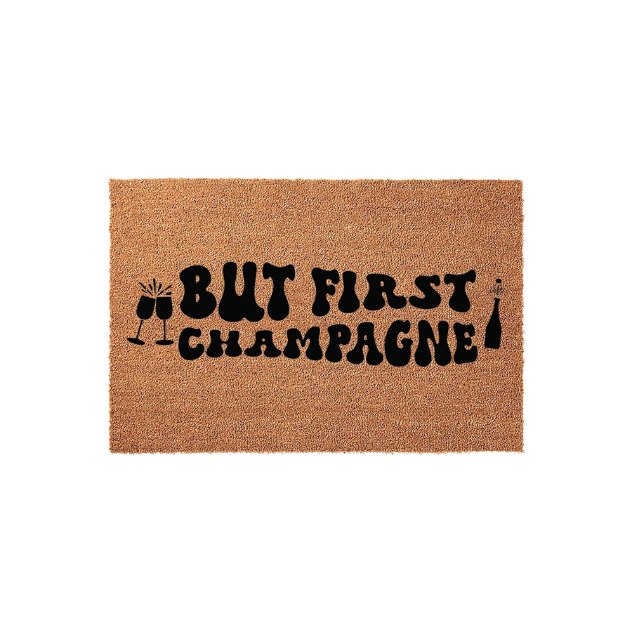 But First Champagne coir doormat featuring a natural brown fiber texture and bold black retro-style lettering, complete with graphic illustrations of two clinking champagne flutes and a popping bottle on a clean white background.