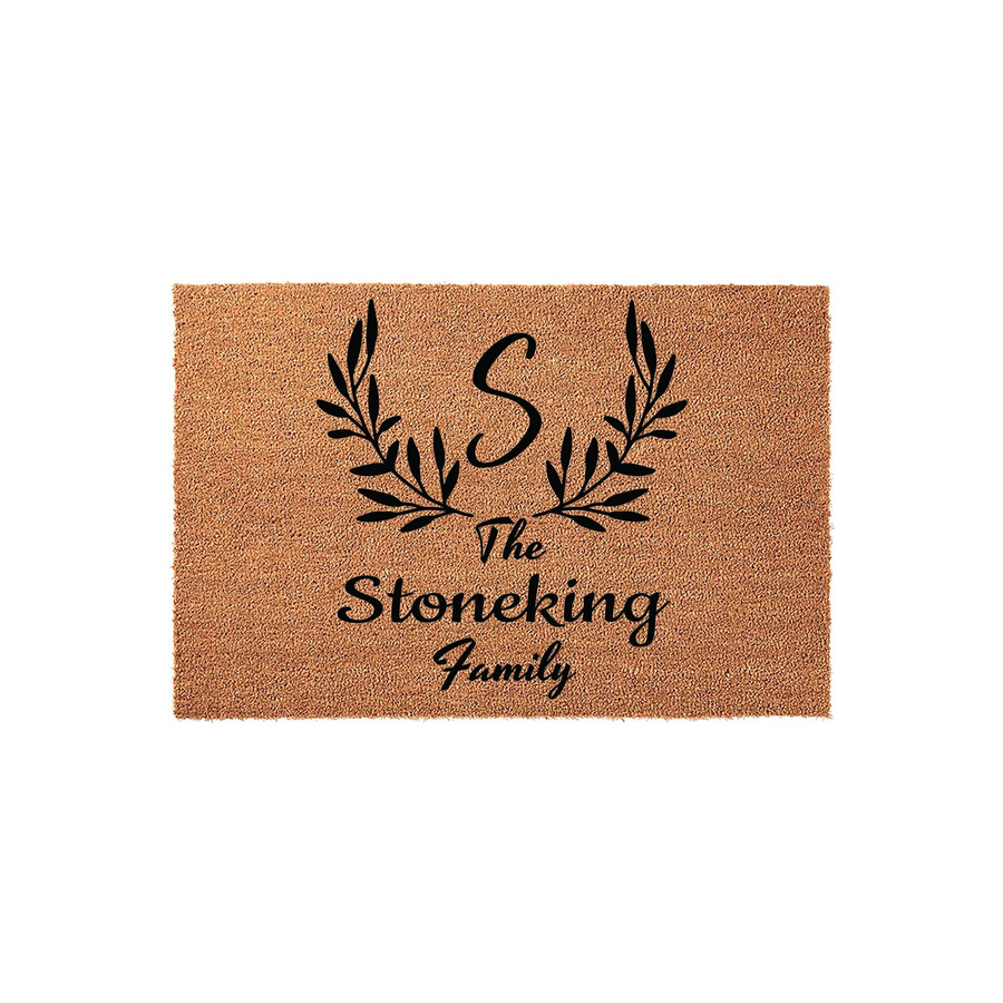 Personalized family name coir doormat featuring a large script initial letter S centered above a decorative black leaf wreath design, creating a welcoming entrance piece for any home with a natural tan fiber texture and black ink.