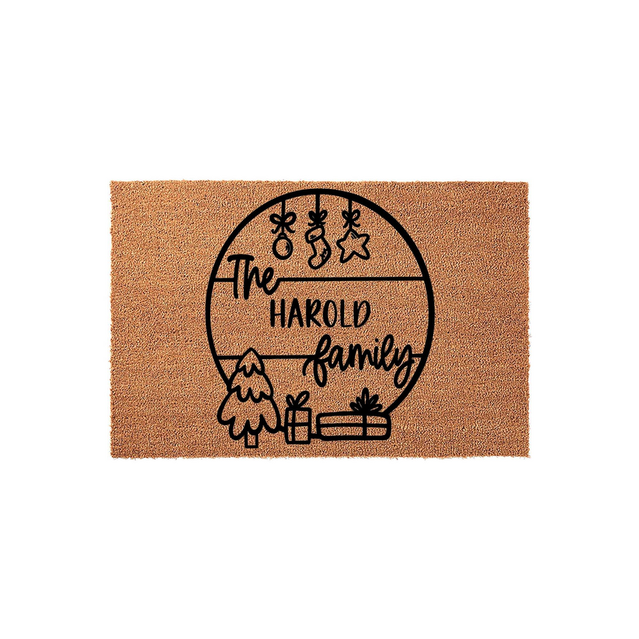 Personalized Christmas family name coir doormat featuring a circular design with hanging ornaments, a small tree, and wrapped gifts, providing a festive welcome for the holiday season on a natural tan textured fiber background.