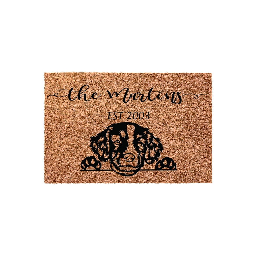 Personalized family name coir doormat featuring a cute illustrated Spaniel dog peeking over the bottom edge, with custom text and an established year printed in black ink on a natural brown textured fiber background.