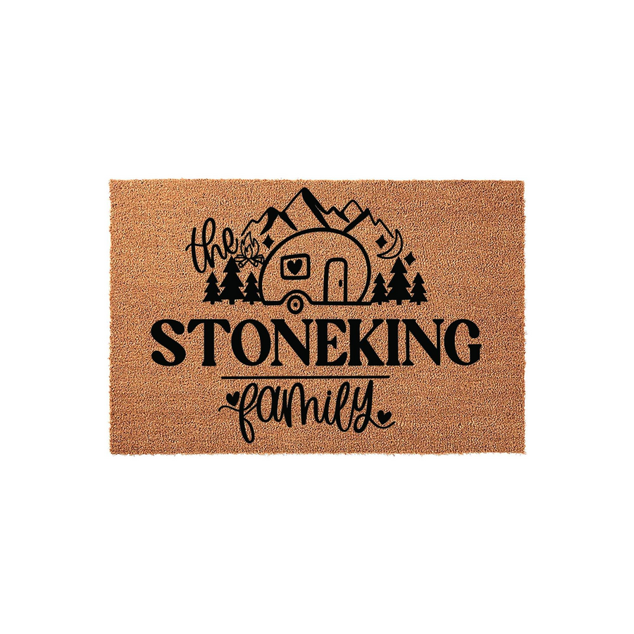 Personalized camping family doormat featuring a black graphic of a camper, mountains, and pine trees on a natural coir background, with space for a custom family name printed in bold lettering above the word family in a script