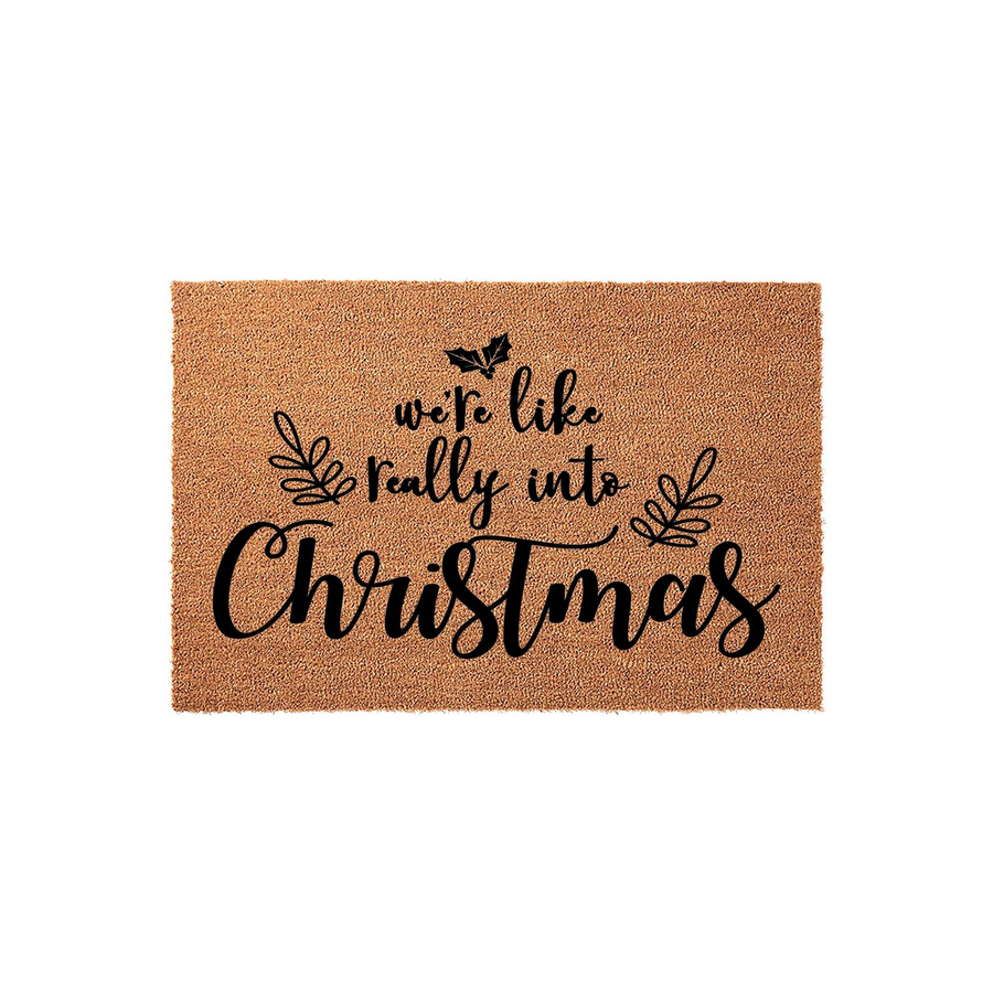 Christmas coir doormat featuring the phrase we are like really into Christmas in elegant black script lettering, accented with decorative holly leaves and sprigs on a natural brown textured fiber background for holiday home decor.