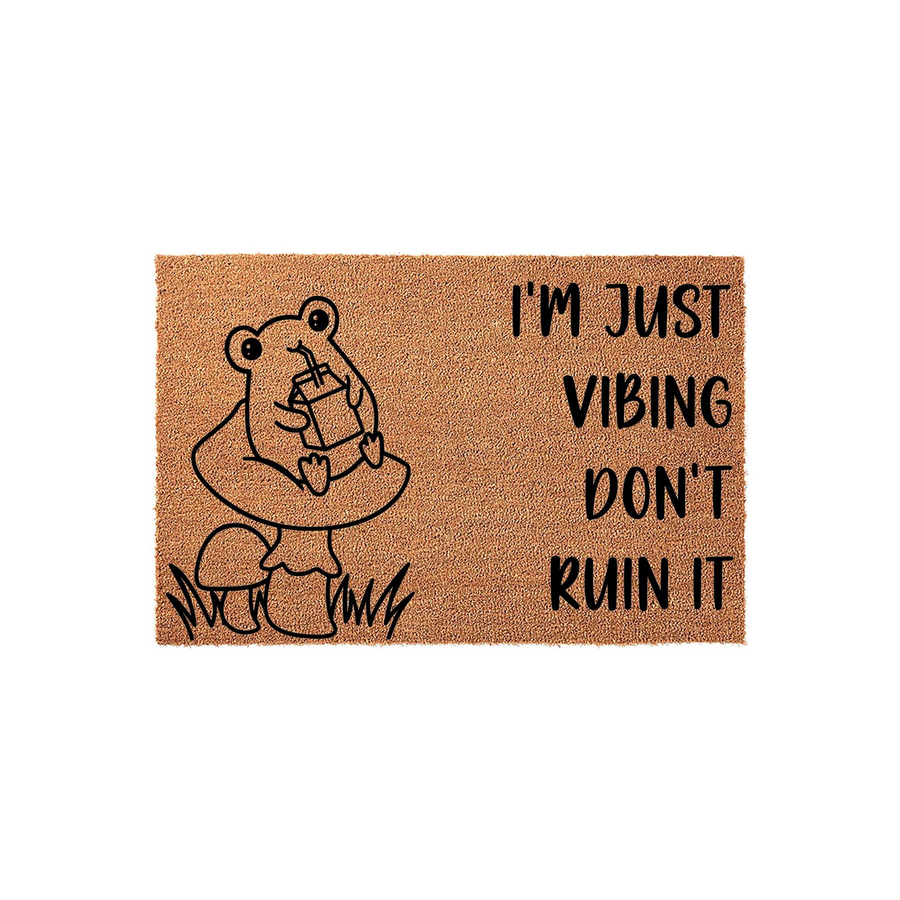 Funny Frog Mushroom Doormat featuring a cute illustration of a frog sitting on a mushroom while drinking from a juice box, accompanied by the text I'm Just Vibing Don't Ruin It printed in black on a natural brown coir fiber