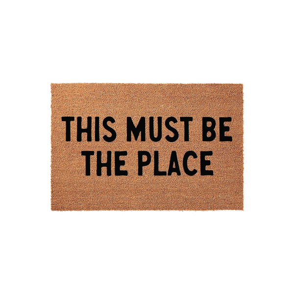 This Must Be The Place coir doormat features a natural brown fiber texture with bold black lettering centered on the mat, creating a welcoming and stylish entrance piece for your front porch or home entryway decor.