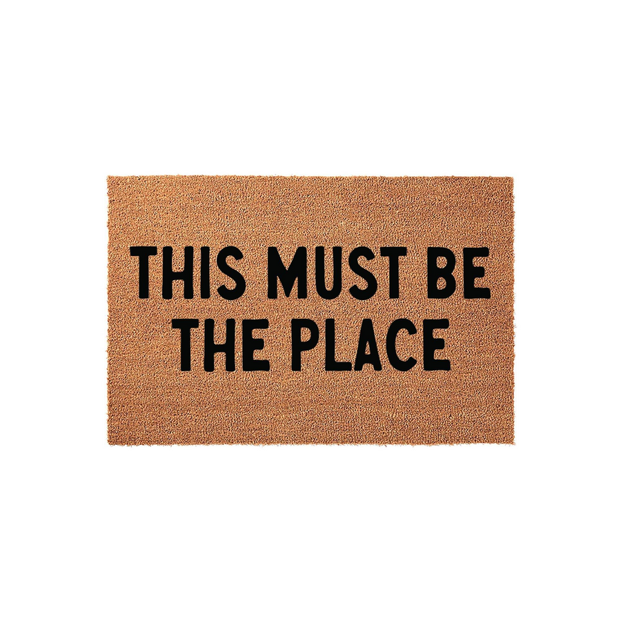 This Must Be The Place coir doormat features a natural brown fiber texture with bold black lettering centered on the mat, creating a welcoming and stylish entrance piece for your front porch or home entryway decor.