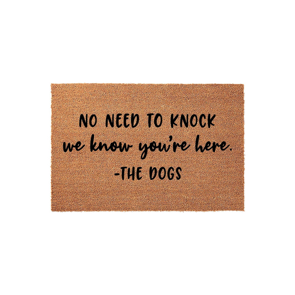 Funny Dog Welcome Doormat featuring a natural coir texture with black printed text that reads No Need To Knock We Know You Are Here, signed by The Dogs, making it a humorous front porch decor piece for any pet lover home entrance.