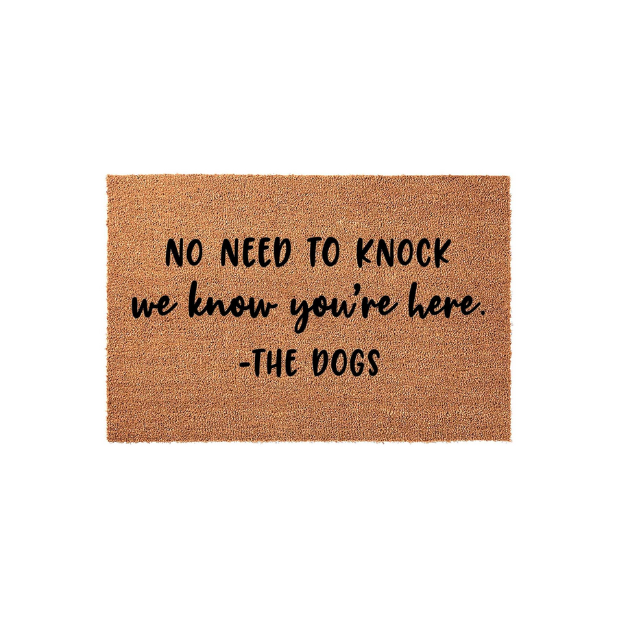 Funny Dog Welcome Doormat featuring a natural coir texture with black printed text that reads No Need To Knock We Know You Are Here, signed by The Dogs, making it a humorous front porch decor piece for any pet lover home entrance.