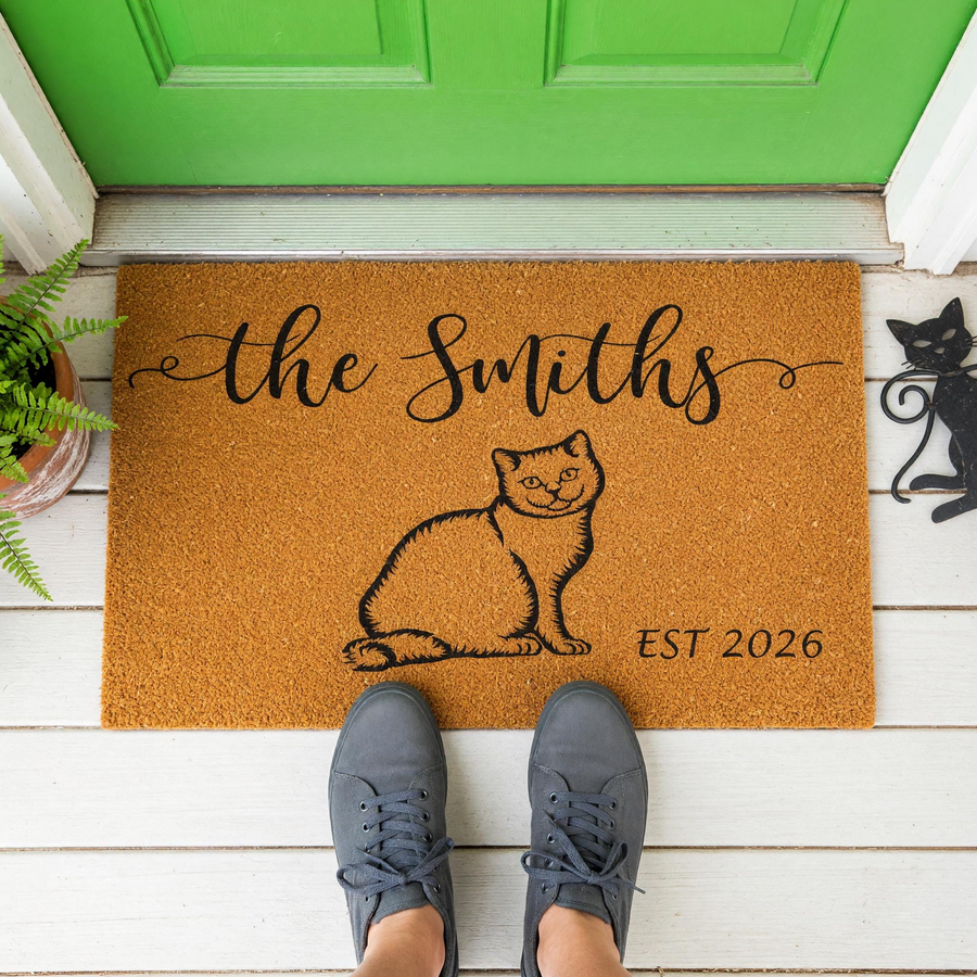 Personalized Cat Family Doormat featuring a cute illustrated cat, custom family name, and established year, displayed on a white porch in front of a bright green door with a person wearing gray sneakers standing on the natural