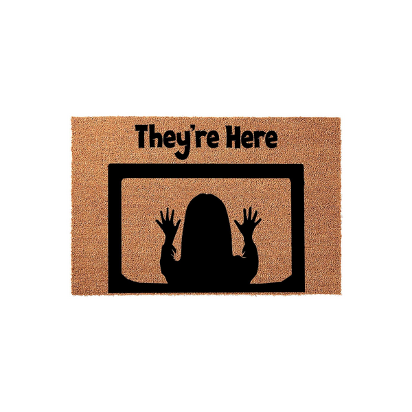 Horror movie themed They're Here coir doormat featuring a black silhouette of a person with hands pressed against a television screen, set against a natural brown textured fiber background for a spooky front porch entryway