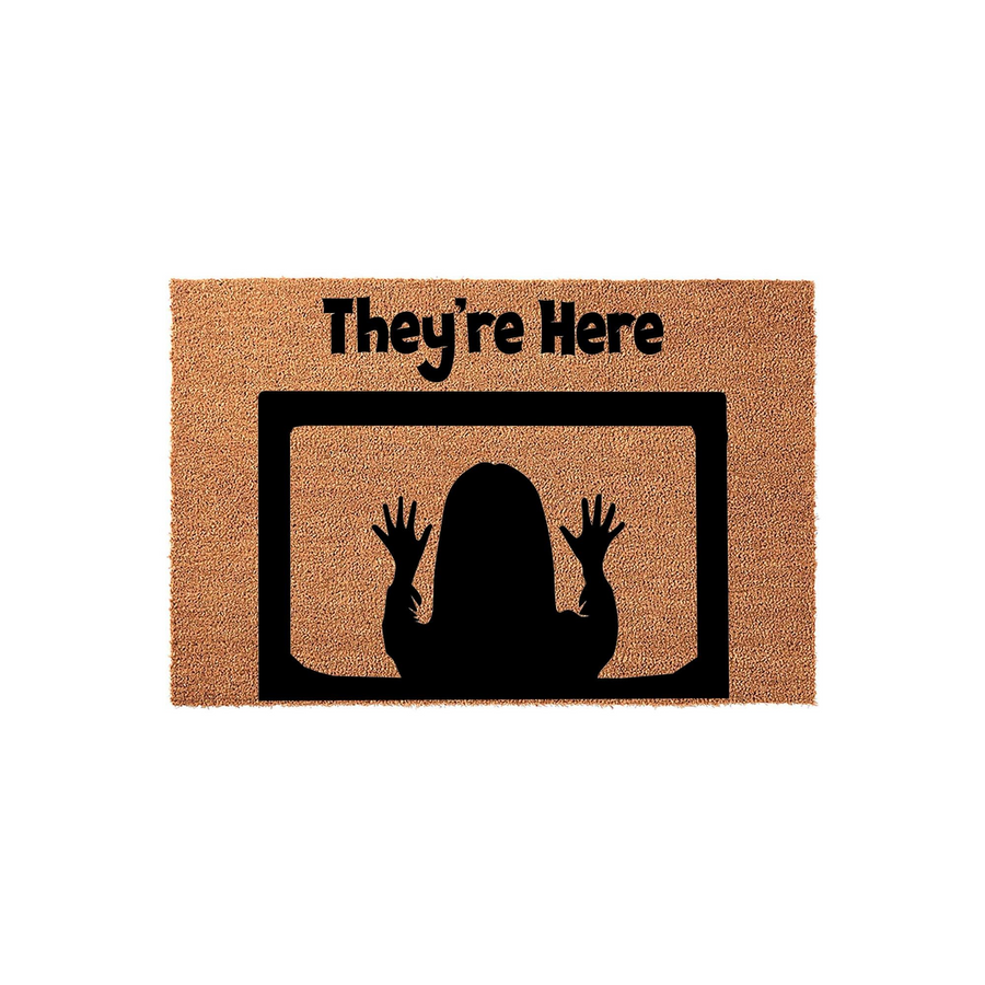Horror movie themed They're Here coir doormat featuring a black silhouette of a person with hands pressed against a television screen, set against a natural brown textured fiber background for a spooky front porch entryway