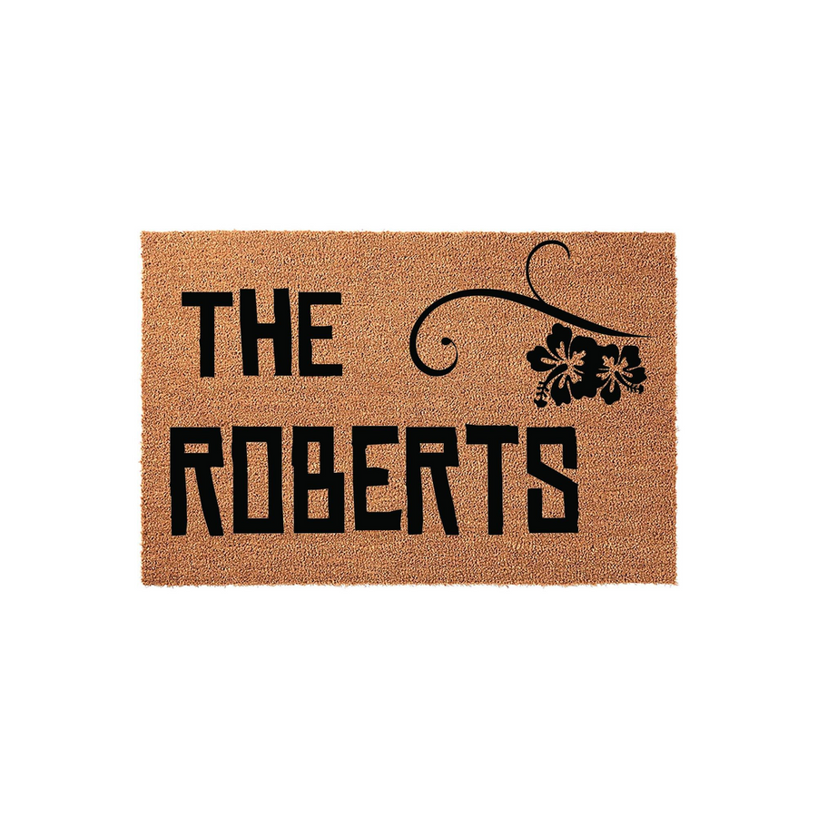 Personalized family name coir doormat featuring a natural tan fiber texture with bold black lettering and a decorative hibiscus flower accent, providing a welcoming touch for any front porch or entryway decor.
