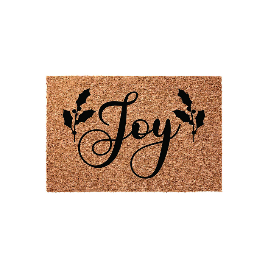 Joy Christmas coir doormat featuring a natural tan fiber texture with black script lettering in the center, flanked by decorative holly leaf illustrations on both sides for a festive holiday entryway decor accent piece.
