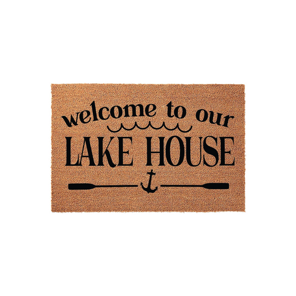 Welcome to our Lake House coir doormat featuring a natural tan fiber texture with black printed text, wavy water lines, and a nautical anchor icon centered between two horizontal oars on a clean white studio background.