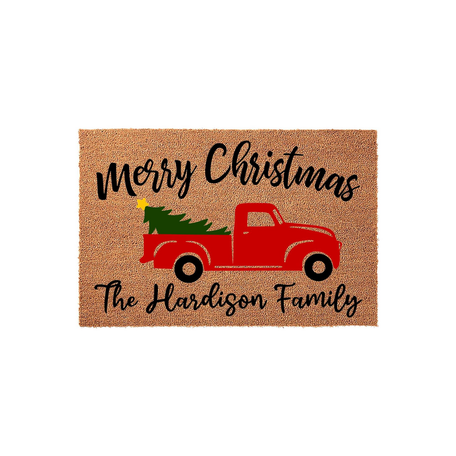 Personalized Merry Christmas coir doormat featuring a festive red truck carrying a green pine tree, with custom family name text printed in black script on a natural tan textured background for a holiday home entryway decor.