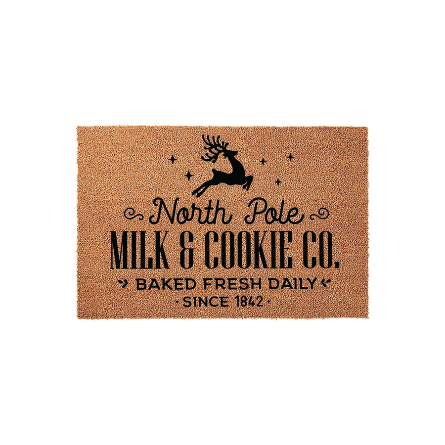 North Pole Milk and Cookie Co. Christmas doormat made of natural coir fiber, featuring a black silhouette of a leaping reindeer, festive stars, and vintage-style typography on a plain white background for holiday home decor.