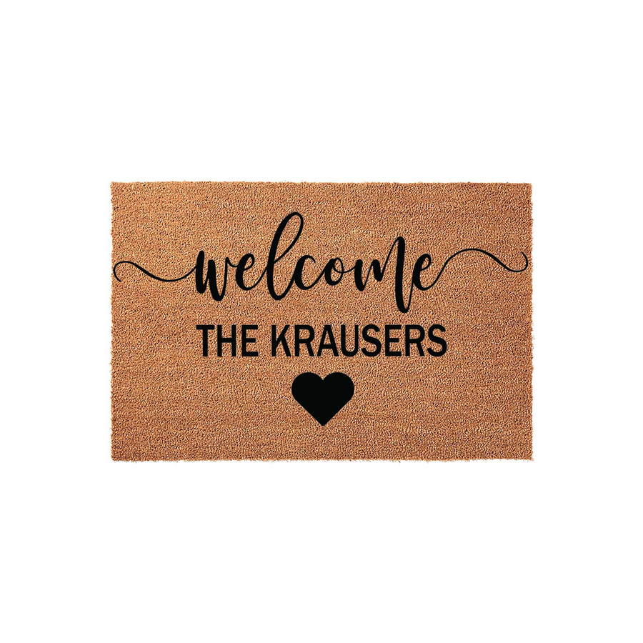 Personalized family name welcome doormat made of natural coir fiber, featuring the word welcome in elegant script, a custom family name in bold black lettering, and a simple heart icon centered on a plain white studio background.