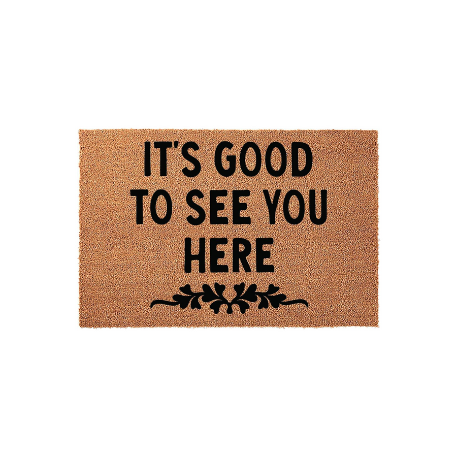 It's Good To See You Here coir doormat featuring a natural tan fiber texture and bold black lettering, accented with a decorative floral flourish at the bottom to create a welcoming entrance for your home front porch or entryway.