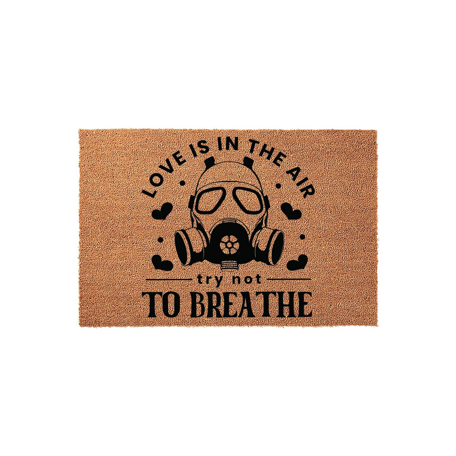 Funny Novelty Doormat featuring a black graphic of a gas mask surrounded by small hearts, with the text Love is in the air try not to breathe printed in bold black lettering on a natural tan coir fiber background for a unique