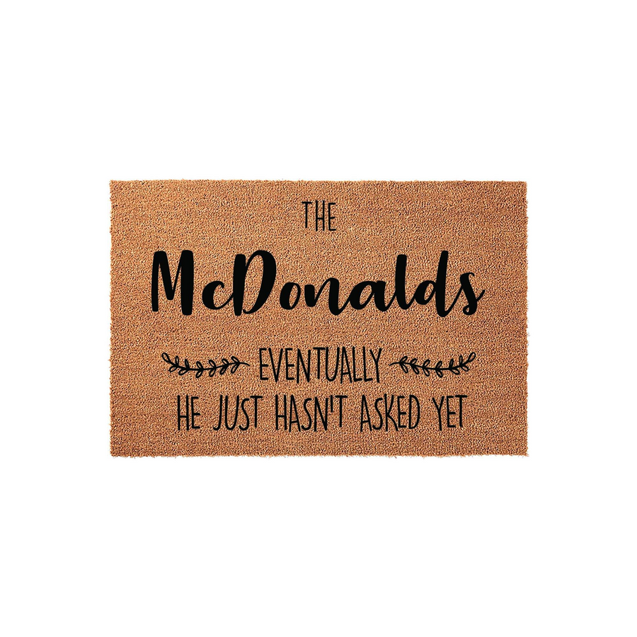 Personalized coir doormat featuring a humorous family name design that reads The McDonalds, Eventually He Just Hasn't Asked Yet, set against a natural brown fiber background with decorative leaf accents for a modern entryway
