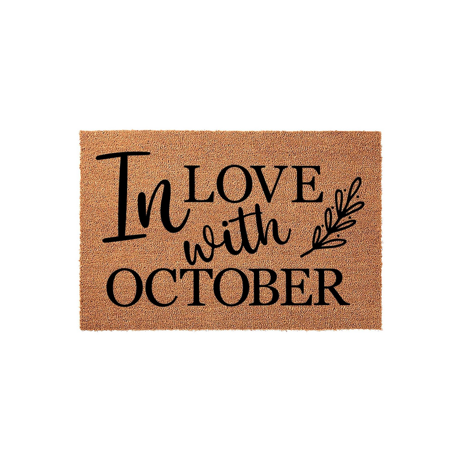In Love with October coir doormat featuring a natural tan fiber texture and bold black lettering that reads In Love with October, accented by a delicate leafy branch design for a festive seasonal front porch decor aesthetic.