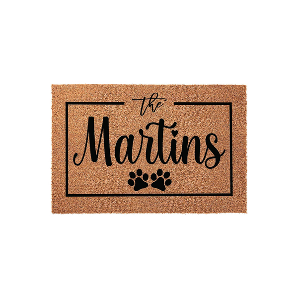 Personalized family name coir doormat featuring a black rectangular border, elegant script text, and two cute paw prints at the bottom, creating a welcoming entrance for any pet-loving home on a plain white studio background.