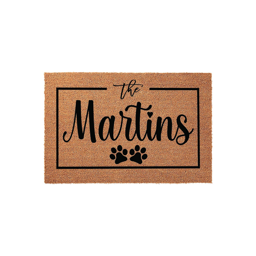 Personalized family name coir doormat featuring a black rectangular border, elegant script text, and two cute paw prints at the bottom, creating a welcoming entrance for any pet-loving home on a plain white studio background.