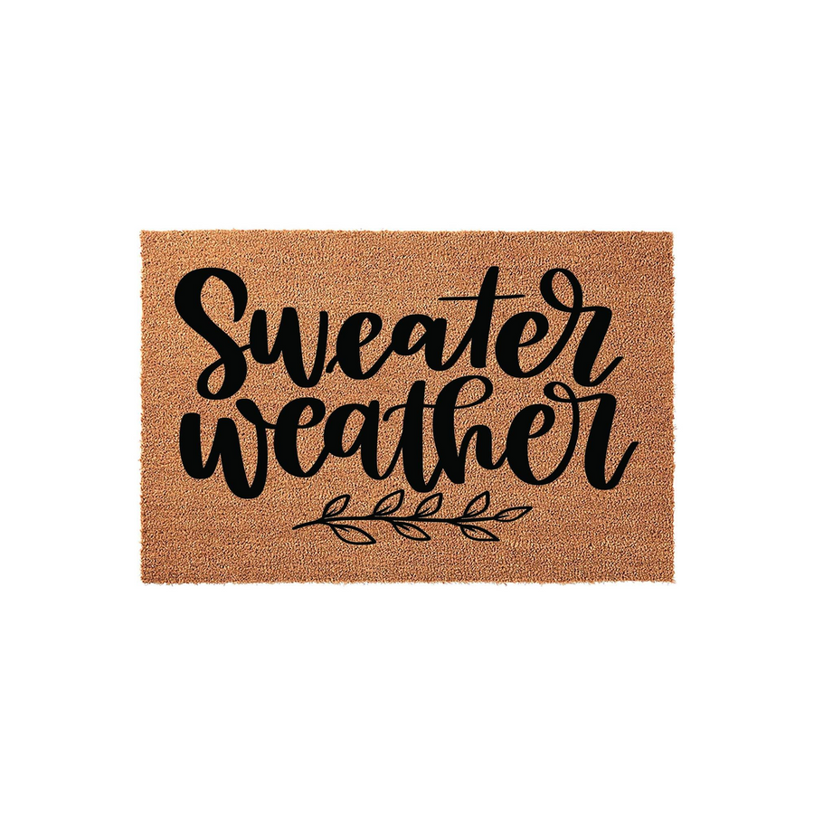 Sweater Weather coir doormat featuring a natural brown fiber texture and bold black script lettering, accented by a simple decorative leaf branch design at the bottom for a cozy fall entryway decor style on a plain white
