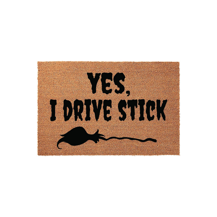 Yes I Drive Stick funny doormat made of natural coir fiber, featuring bold black text and a silhouette of a witch flying on a broomstick, perfect for a whimsical front porch entrance that showcases a love for Halloween and