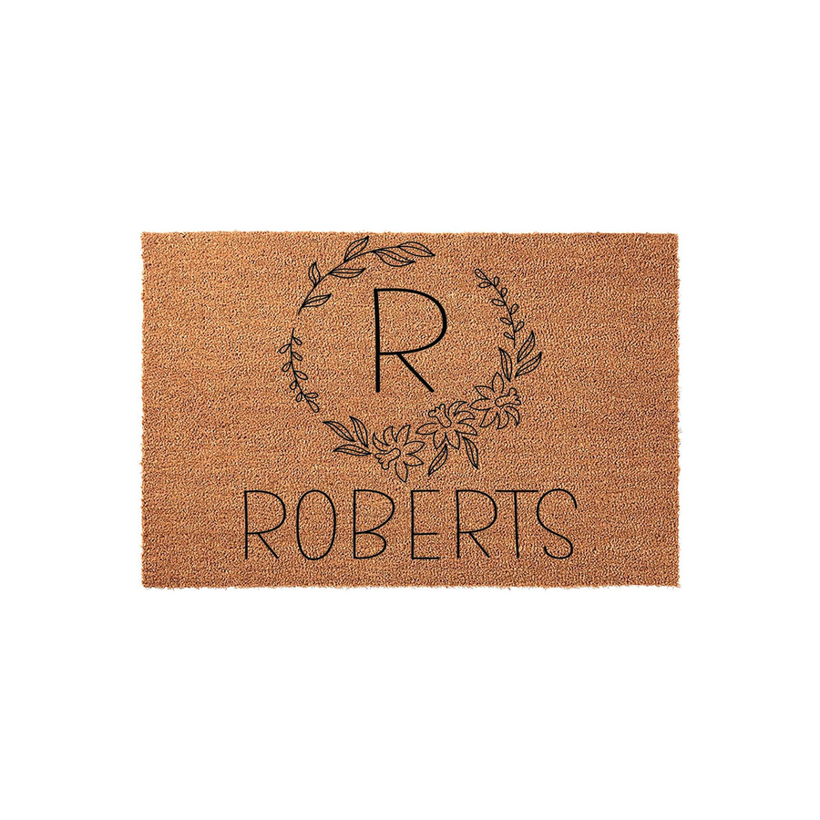 Personalized coir doormat featuring a custom last name and a large initial letter centered within a delicate black line art floral wreath, creating a welcoming entryway decor piece for a new home or a housewarming celebration.