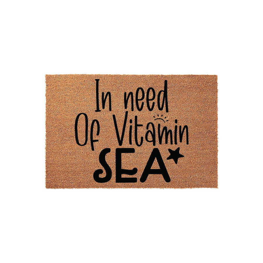 In Need Of Vitamin Sea Coir Doormat featuring a natural brown fiber texture with bold black lettering that reads In Need Of Vitamin SEA with a small sun and star icon, perfect for a beach house or coastal home entryway decor.