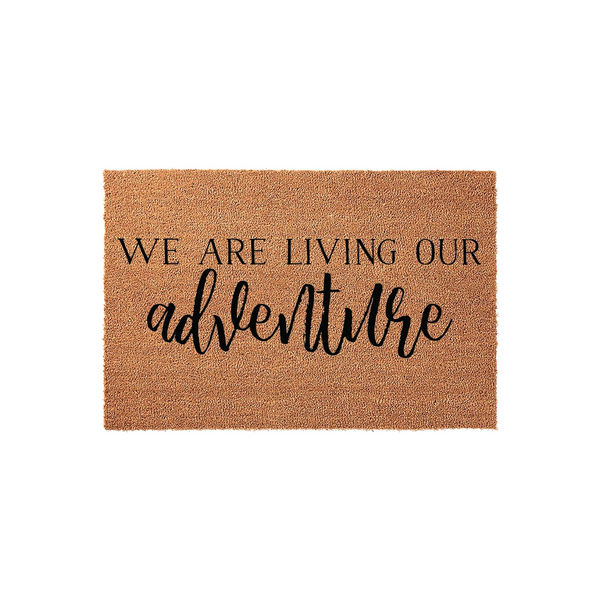 We Are Living Our Adventure Coir Doormat featuring a natural tan fiber texture and bold black lettering, providing a rustic and welcoming entrance decor piece for any home front porch or entryway floor space.