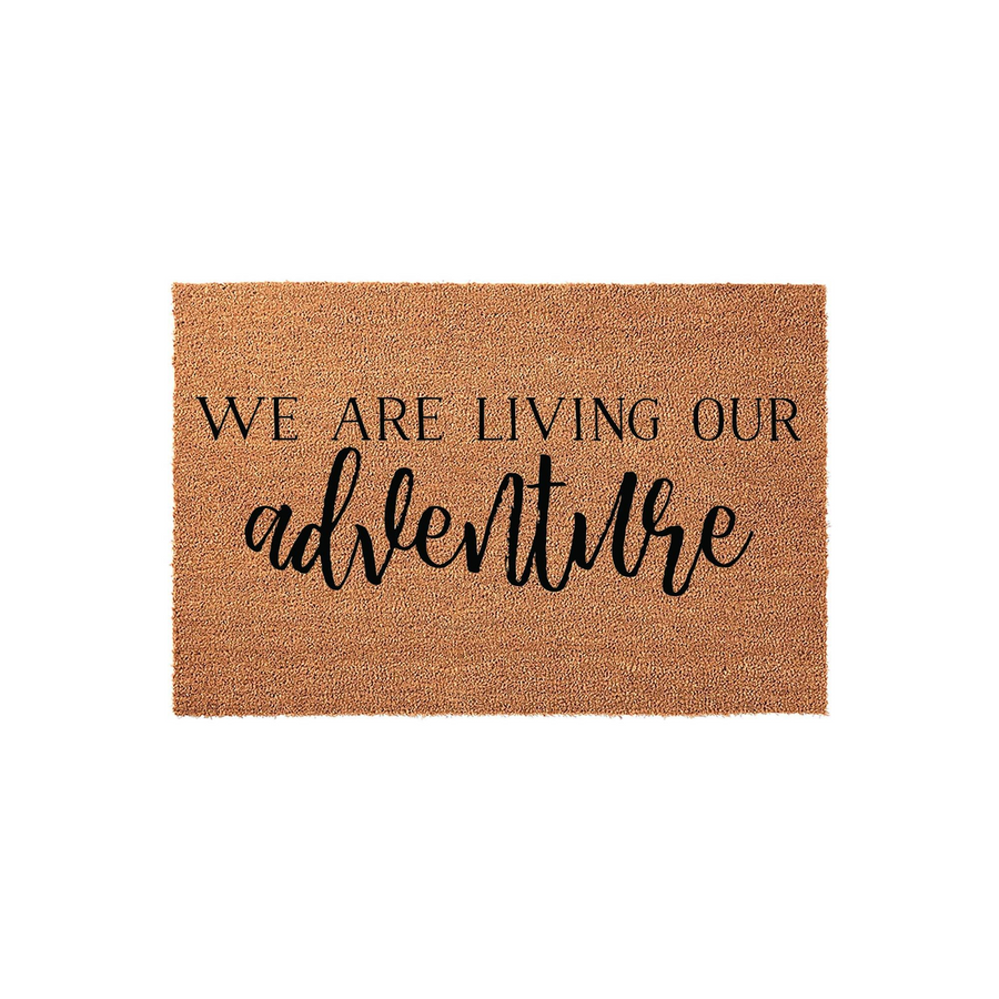 We Are Living Our Adventure Coir Doormat featuring a natural tan fiber texture and bold black lettering, providing a rustic and welcoming entrance decor piece for any home front porch or entryway floor space.