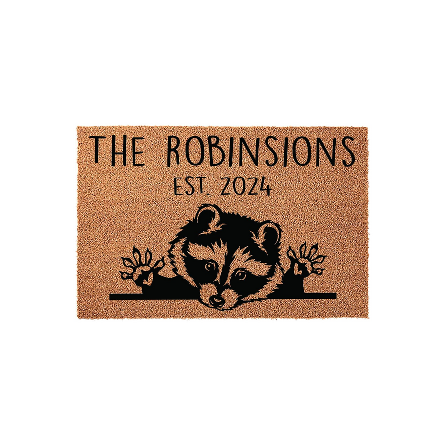 Personalized Raccoon Welcome Doormat featuring a cute black illustration of a raccoon peeking over a line, with custom family name text and established year printed on a natural brown coir fiber mat against a clean white
