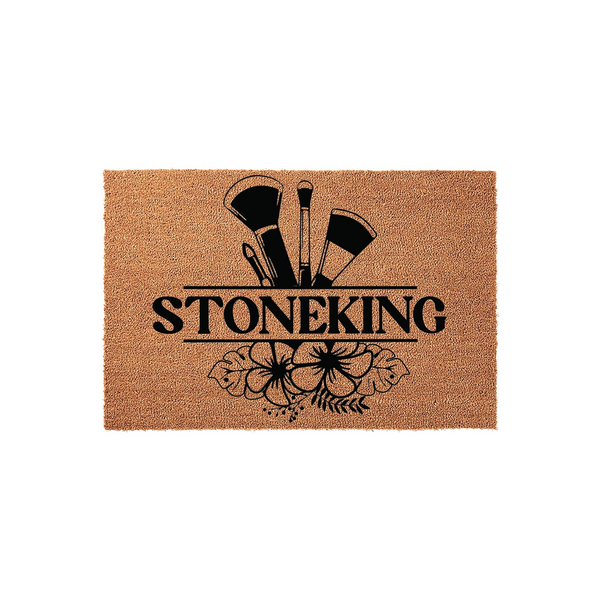 Personalized makeup artist coir doormat featuring a black graphic of professional cosmetic brushes and tropical hibiscus flowers, with a central horizontal line for a custom family name on a natural tan textured fiber background.
