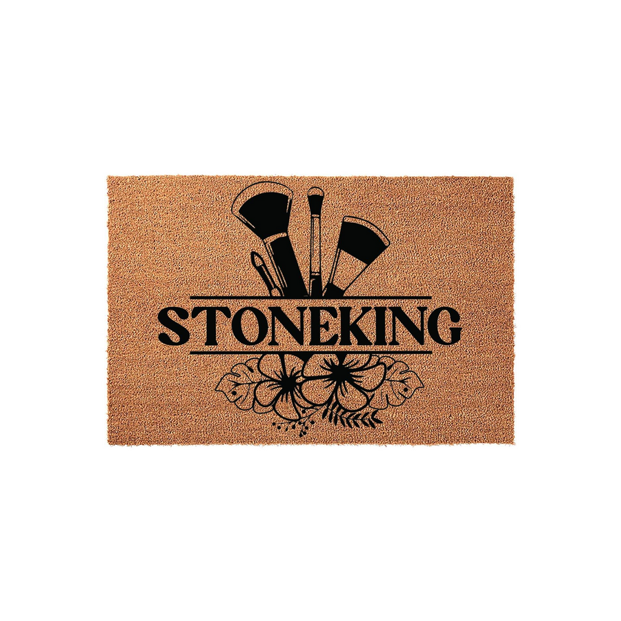 Personalized makeup artist coir doormat featuring a black graphic of professional cosmetic brushes and tropical hibiscus flowers, with a central horizontal line for a custom family name on a natural tan textured fiber background.