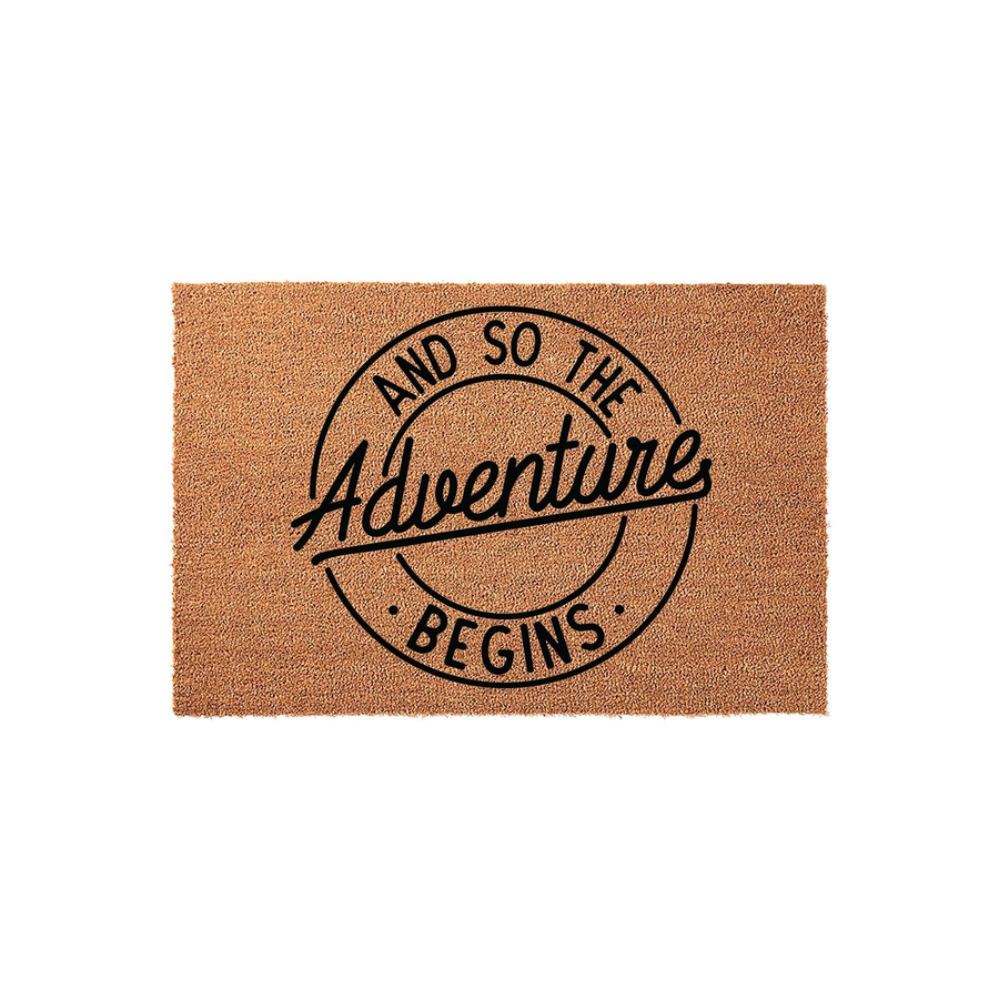 And So The Adventure Begins coir doormat featuring a circular black graphic design printed on a natural tan fiber surface, providing a rustic and welcoming entrance decor piece for a new home or apartment entryway.