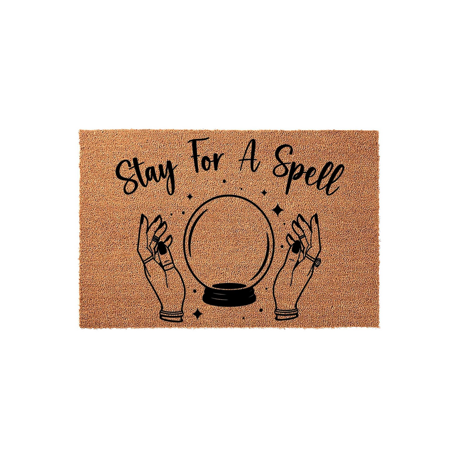 Stay For A Spell Halloween doormat featuring a natural coir texture with a black printed design of two hands holding a crystal ball, creating a spooky and festive entryway decor piece for the fall season on a plain white