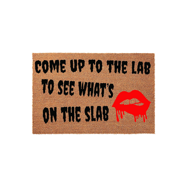 Rocky Horror Picture Show themed coir doormat featuring the quote Come Up To The Lab To See What Is On The Slab in bold black lettering next to a vibrant red graphic of dripping lips on a natural brown textured fiber background.