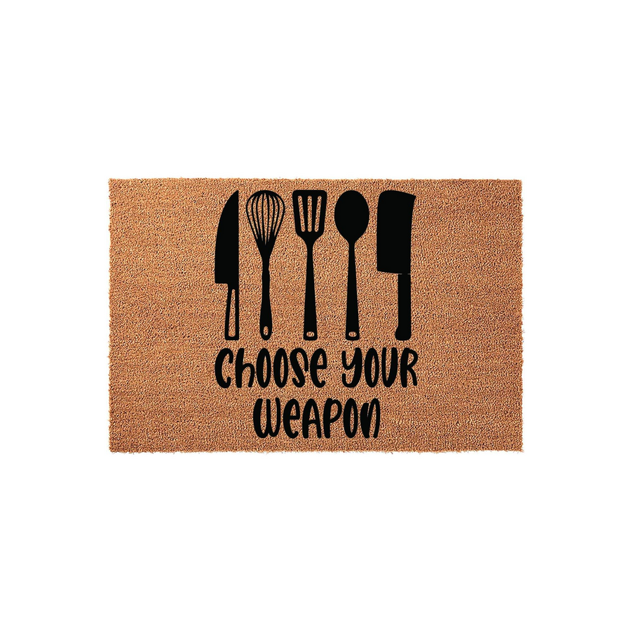 Choose Your Weapon kitchen doormat made of natural coir fiber, featuring a black printed design of five cooking utensils including a knife, whisk, spatula, spoon, and cleaver centered on a rectangular tan textured background.