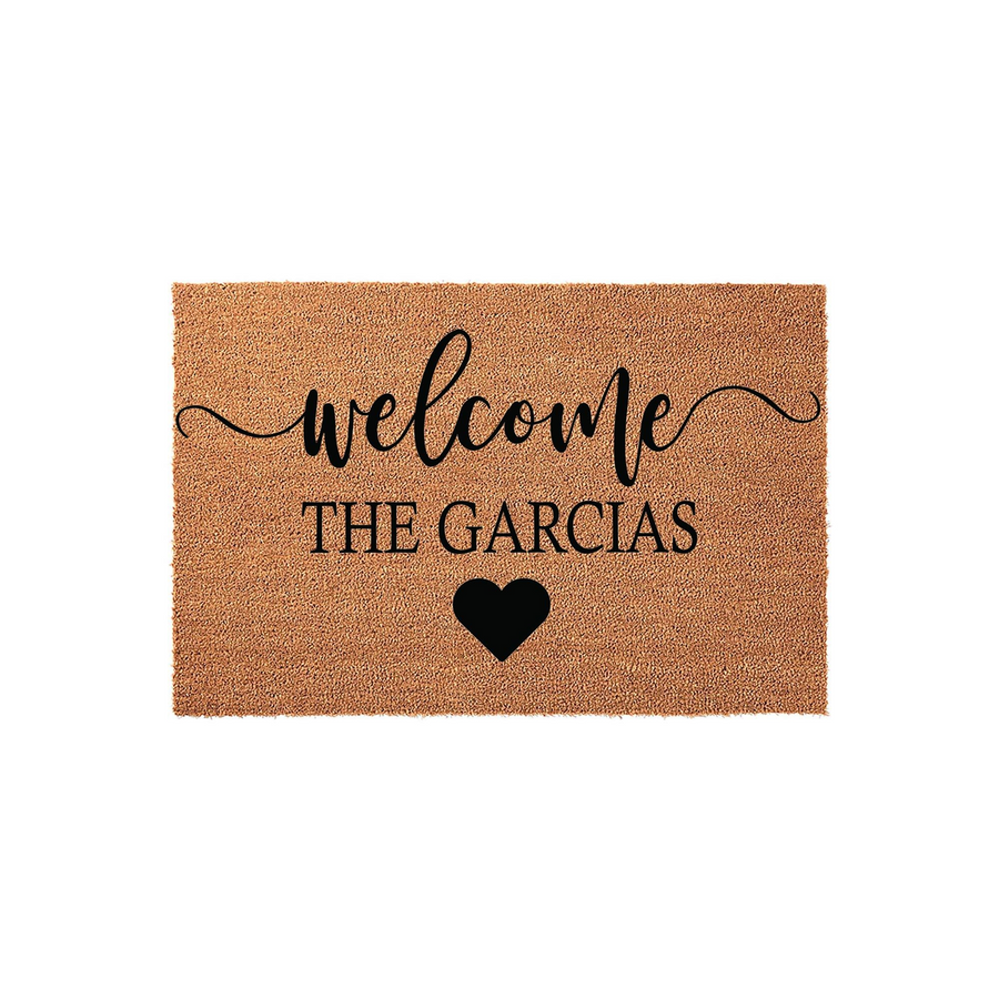 Personalized coir welcome doormat featuring the word welcome in elegant script, a custom family name line, and a simple black heart icon centered on a natural brown textured fiber background for a classic entryway decor look.